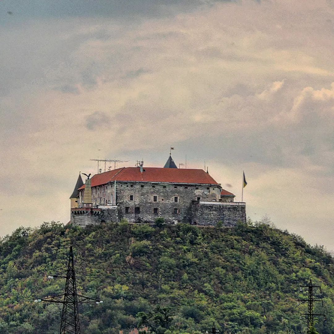 Várkastély kőfalakkal és piros tetővel egy zöld dombtetőn, felhős ég alatt.
